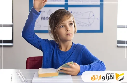 تُظهر طفلاً يرتدي قميصًا أزرق، جالسًا في فصل دراسي ورافعًا يده للإجابة على سؤال. يبدو أنه يطلب دعمًا أكاديميًا أو يشارك بفاعلية في الدرس، وهو يحمل كتابًا مفتوحًا، مما يؤكد اهتمامه بالتعلم