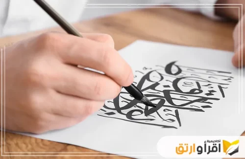 تُظهر الصورة يد شخص يكتب فن كتب خط عربي​ باستخدام قلم خاص على ورقة بيضاء، حيث تبدو الحروف مرسومة بدقة وعناية، مع حضور كبير لجمالية الحروف العربية