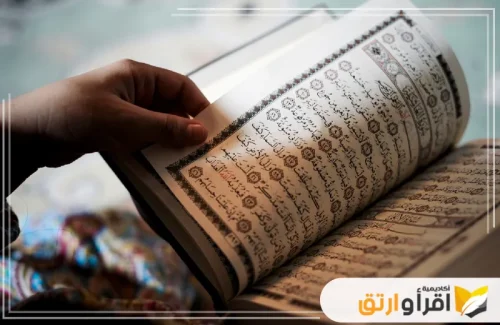 يدًا تمسك بكتاب القرآن الكريم، مع التركيز على صفحاته المكتوبة بخط جميل وواضح. تظهر بعض حلقات السند على حواف الصفحات، مما يضيف جمالية للطباعة