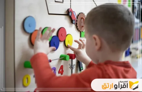 طفلاً صغيراً يتفاعل مع لوح تعليمي مزود بمجموعة من الأشكال والألوان المختلفة، حيث أن دورات تعليم مهارات التواصل بتركيب القطع الملونة في أماكنها. تعكس الصورة بيئة تعليمية مليئة بالأنشطة