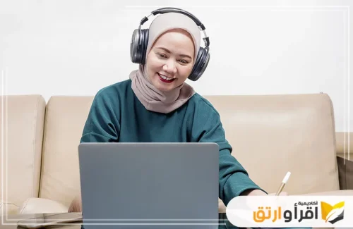  امرأة شابة ترتدي الحجاب وتضع سماعات رأس، وهي تجلس أمام جهاز كمبيوتر محمول وتكتب في دفتر. تعكس هذه اللقطة مدى أهمية التعلم عن بعد في تعزيز التحصيل الدراسي