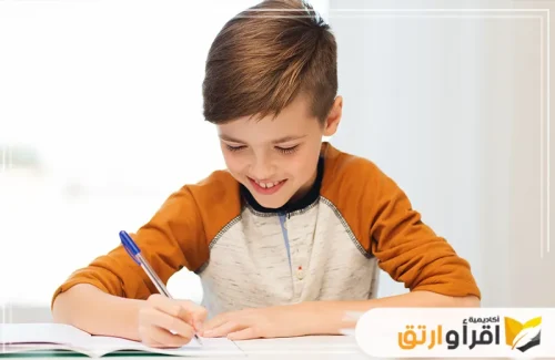 طفلًا صغيرًا يجلس على طاولة وهو يكتب في دفتره مبتسمًا، مما يدل على استمتاعه بما يفعله. يبدو أنه يركز على تحسين الخط العربي.