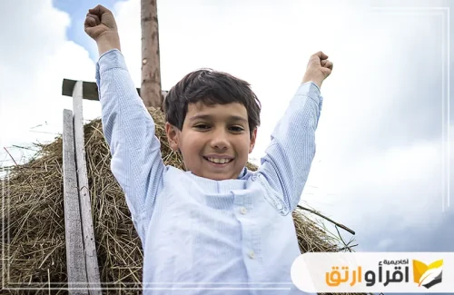 طفلًا مبتسمًا ويرتدي قميصًا أبيض، رافعًا يديه إلى الأعلى وكأنه يشعر بالنجاح والإنجاز. البيئة المحيطة تبدو طبيعية، مع زيادة الثقة بالنفس