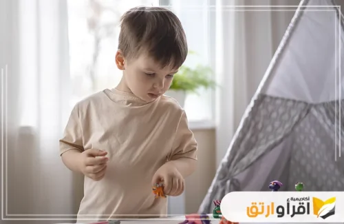 طفلًا صغيرًا، يرتدي قميصًا بلون البيج، وهو يلعب بأشكال صغيرة على سطح أبيض، ويبدو مندمجًا. لعل هذا النشاط يُشبه بعض الأساليب المستخدمة ضمن طرق علاج التوحد عند الأطفال