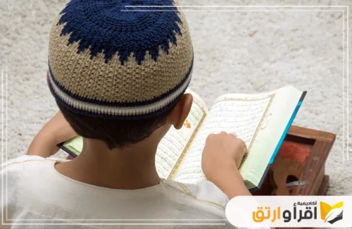 طفلًا صغيرًا وهو يقرأ القرآن الكريم، جالسًا بهدوء مع ارتدائه لقميص أبيض وطاقية تقليدية. يُظهر الطفل اهتمامه وتركيزه في تحفيظ القرآن