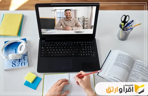 لقطة من الأعلى لشخص يكتب في دفتر ملاحظات أثناء متابعة حصة دراسية عبر الإنترنت على جهاز كمبيوتر محمول. يظهر على الشاشة معلم مبتسم، مما يبرز كيف أصبحت التكنولوجيا والتعليم متكاملين