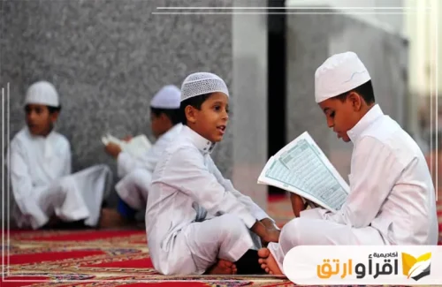 من الأطفال في ملابس بيضاء، يجلسون على سجادة صلاة حمراء. اثنان منهم يتحدثان بينما يقيم الآخرون في أفضل مركز تحفيظ القرآن للأطفال والكبار.