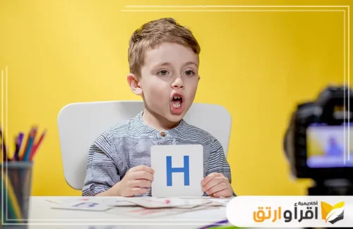 في الصورة، يظهر طفل صغير يجلس أمام طاولة، ممسكًا ببطاقة تحمل حرف، وهو يوجه نظره نحو الكاميرا أثناء نطقه للحرف. علاج تاخر النطق عند الاطفال​