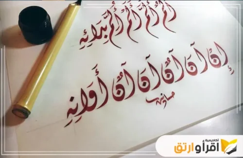 عملًا فنيًا بالخط العربي مكتوبًا بحبر أحمر وبقلم تقليدي، مع زجاجة الحبر بجانب الورقة، مما يجسد جمال فنون خط عربي منقط. يتضح أن الصورة أُخذت في سياق تعليمي وتدريبي